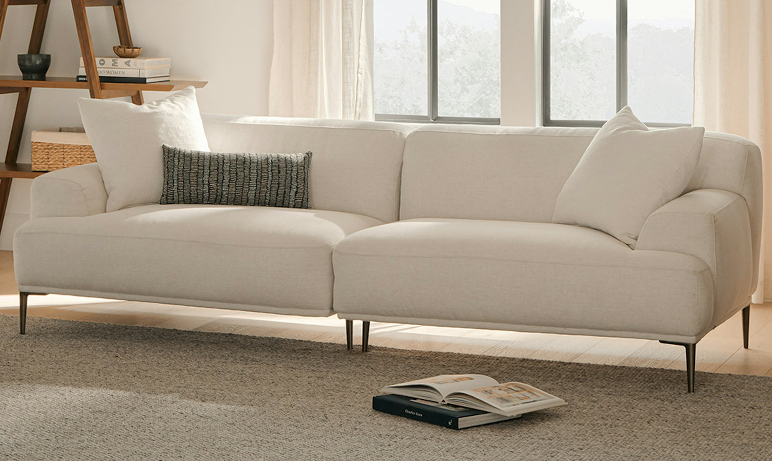 A modern living room with a white sofa adorned with pillows, situated on a neutral-toned rug. A book lies open on the floor in front of the sofa. Light filters in through large windows in the background, highlighting a corner shelf with decorative items on the left.