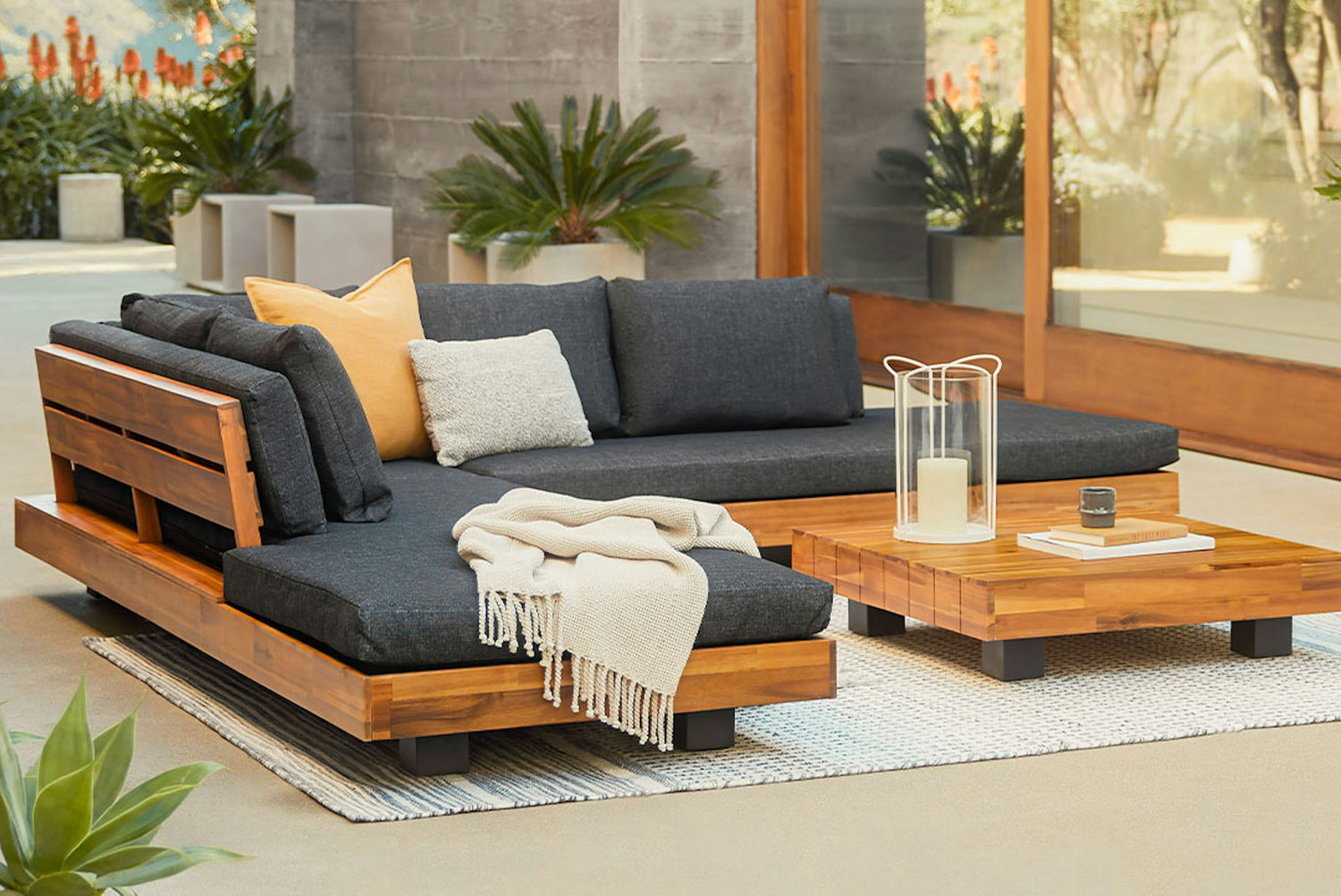A modern outdoor patio setup featuring a wooden sectional sofa with dark cushions, adorned with gray and yellow throw pillows and a beige blanket. A matching wooden coffee table holds a candle lantern and a small decorative item, with plants in the background.