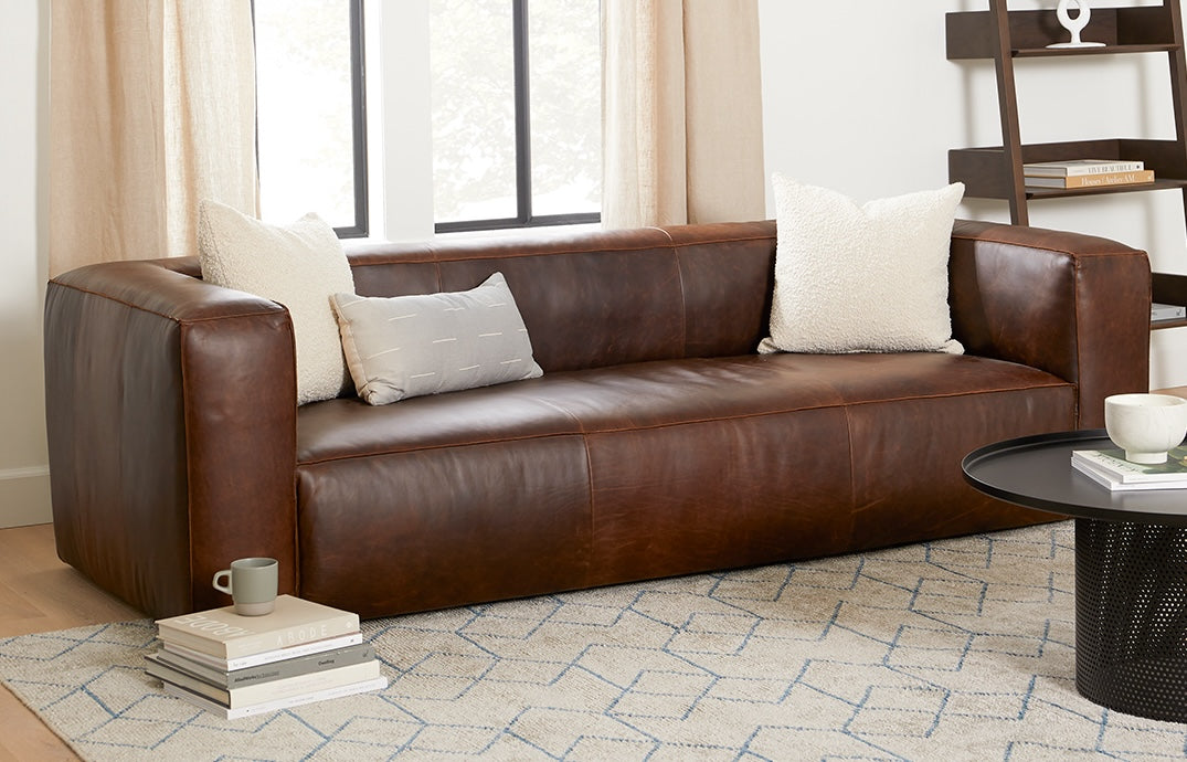 A brown leather sofa with two white pillows and one gray pillow is set against a backdrop of beige curtains and large windows. In front of the sofa, there is a round black coffee table with a cup and several books. The floor is covered with a light-colored rug with a blue geometric pattern. Some stacked books with a coffee cup sit to the left.