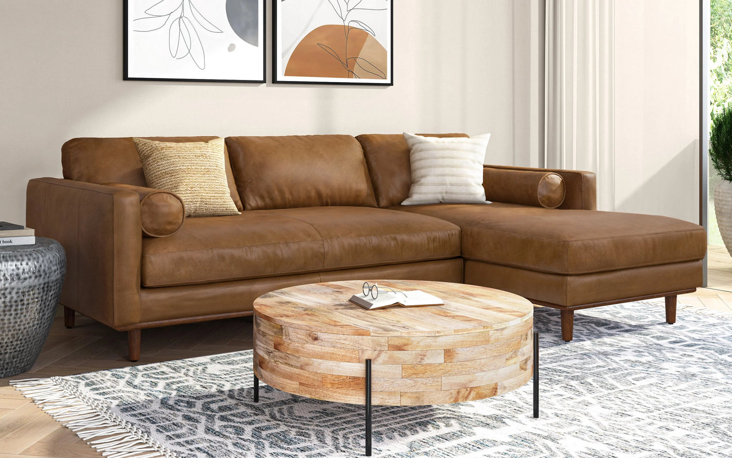 A cozy living room features the Morrison Mid Century Sectional in Genuine Leather, complemented by two pillows and a round wooden coffee table. Abstract art adorns the wall, while a potted plant adds life to the background, creating a light and airy ambiance in the room.