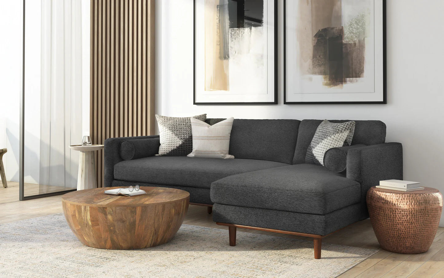 A modern living room featuring the Morrison Mid Century Sectional in Genuine Leather, accented with patterned pillows. A round wooden coffee table and a copper side table rest on a light textured rug. Two abstract paintings grace the wall, complemented by a large window with sheer curtains.