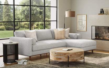 A modern living room featuring the Morrison Mid Century Sectional in genuine leather, complemented by a round wooden coffee table, floor lamp, and abstract wall art. Large windows provide a garden view. The room includes a fireplace and maintains a neutral color palette.