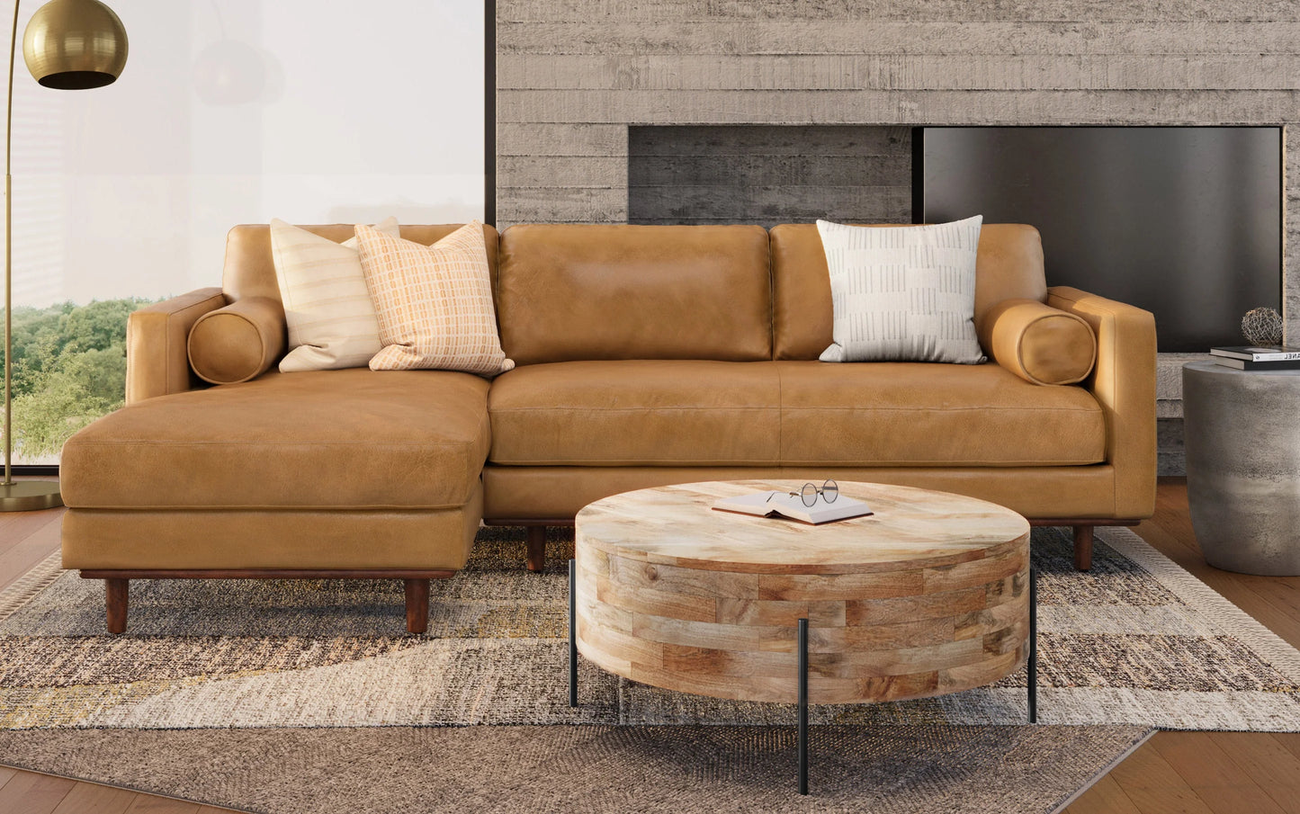 A cozy living room showcases the Morrison Mid Century Sectional in Genuine Leather, embellished with three patterned throw pillows. A round wooden coffee table, holding a book and glasses, rests on a textured rug. The ambiance is enhanced by a modern concrete wall and a window offering a lush green view.