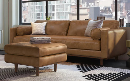 A Morrison 89-Inch Sofa and Ottoman Set in Genuine Leather, complete with two white and patterned cushions, is placed against a large window offering a city view. In front of the sofa, the matching leather ottoman holds a stack of books. The room is complemented by a modern black-and-white patterned rug.