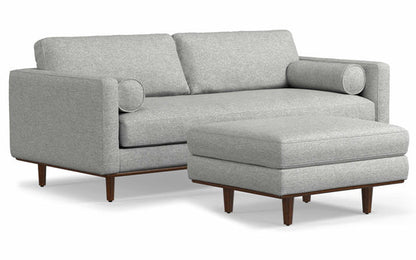 A contemporary Morrison 89-inch sofa with squared arms and twin back cushions, paired with a matching ottoman. Both items are crafted from genuine leather and rest on wooden legs, offering a mid-century design appeal.