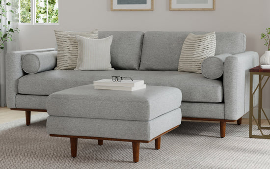 A contemporary living room showcases a Morrison 89-Inch Sofa and Ottoman Set in genuine leather, accompanied by two striped pillows and bolster cushions. The matching ottoman holds a pair of glasses and a book on top. The space is adorned with neutral tones, framed wall art, and is bathed in natural light from the nearby window.