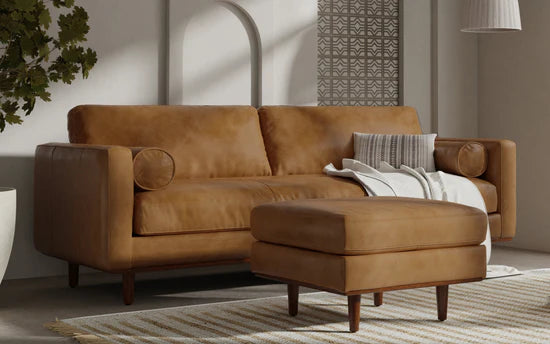 A modern living room featuring the Morrison 89-Inch Sofa and Ottoman Set in genuine leather. A white and gray throw blanket is elegantly draped over the sofa, complemented by a striped pillow. The cozy setting is enhanced by a patterned rug and a plant in a white pot.