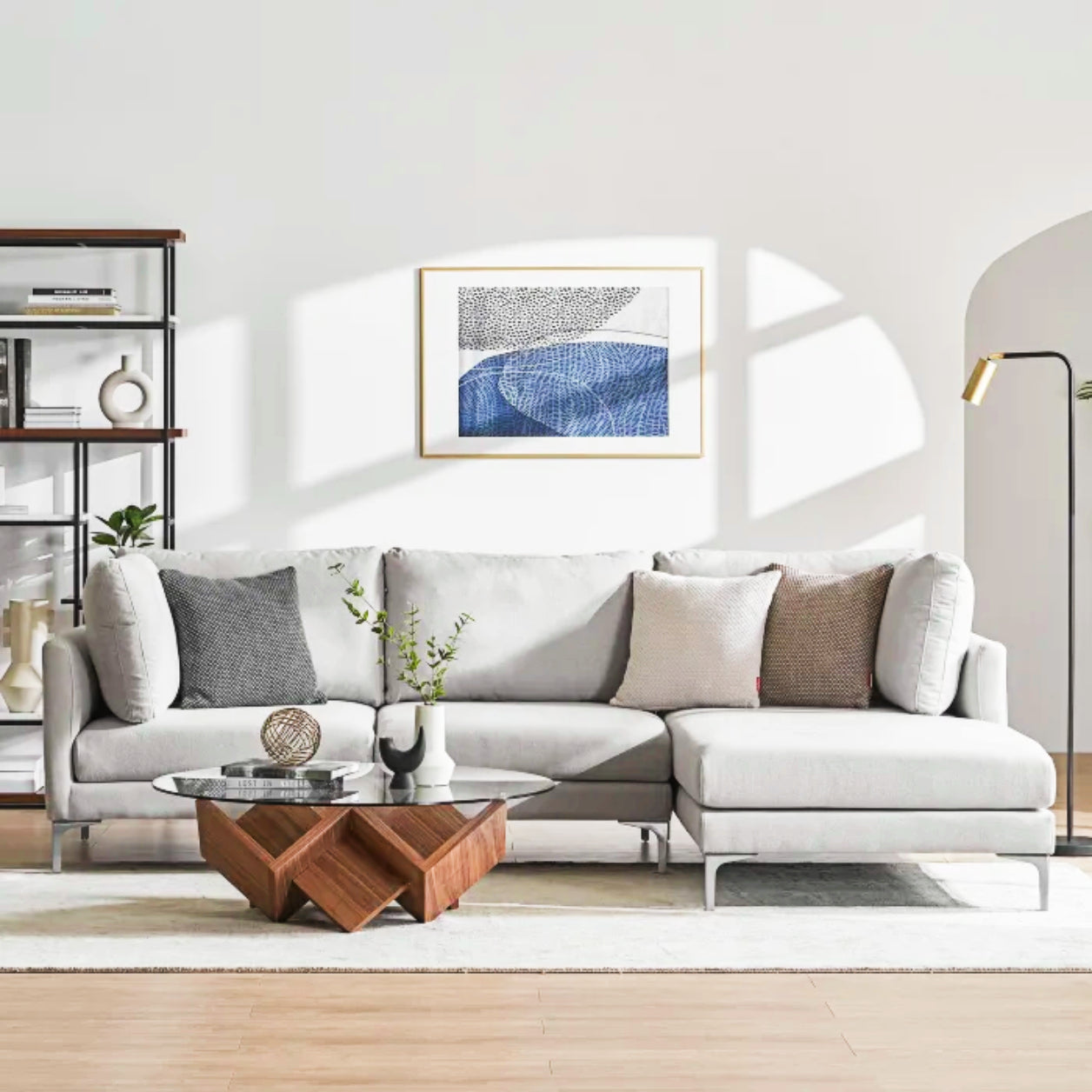 A modern living room featuring the light gray Adams Chaise Sectional Sofa by Castlery is adorned with various cushions. A geometric wooden coffee table with a round glass top is positioned in front. Above hangs a framed abstract art piece, while to the left stands a black shelving unit.