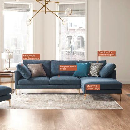 A contemporary living room featuring the Adams Chaise Sectional Sofa by Castlery in dark blue. The couch includes labeled features such as removable cushion covers, deep seats, added support cushions, and sturdy legs. The space is accentuated by large windows, wooden floors, and a rug.