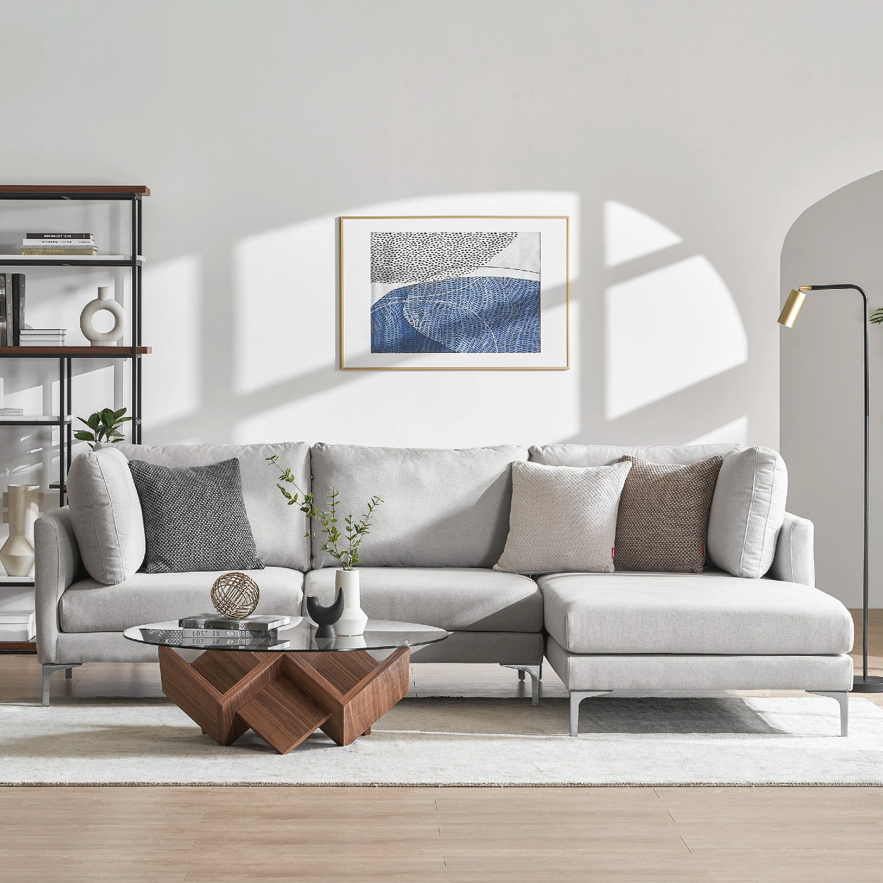 A modern living room showcases the Adams Chaise Sectional Sofa in light gray by Castlery, enhanced with assorted pillows. The space is complemented by a geometric wooden coffee table and a stylish floor lamp. A framed abstract artwork adorns the wall, while shelves display books and decorative items to the side. Natural light brightens the entire room.