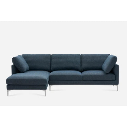 A modern dark blue L-shaped sectional sofa with three back cushions and a chaise on the left. The sofa features sleek silver legs and includes two cylindrical bolster pillows. The design is minimalistic against a white background.