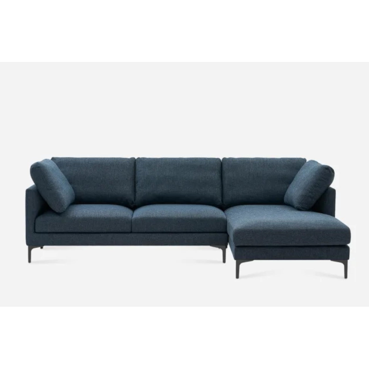 A modern L-shaped dark blue sofa with three seat cushions on the left and a chaise lounge extension on the right. The sofa has four back cushions and two side cushions, supported by slim, black legs against a plain white background.