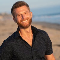 A smiling man with short brown hair and a beard stands on a beach during sunset. He is wearing a black short-sleeved shirt with small white dots. The ocean and sandy shore are in the background, creating a warm and relaxed atmosphere.
