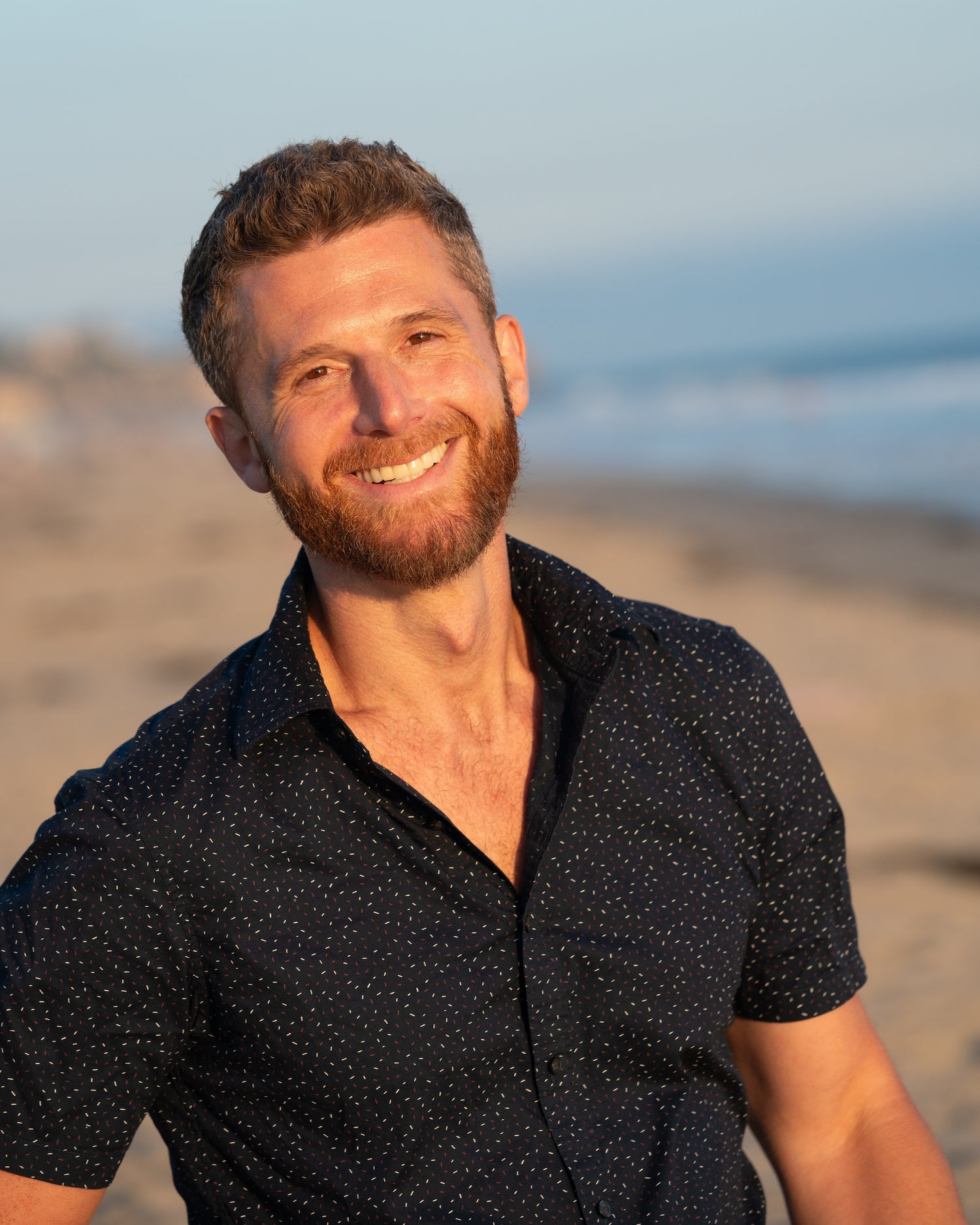 A smiling man with short brown hair and a beard stands on a beach during sunset. He is wearing a black short-sleeved shirt with small white dots. The ocean and sandy shore are in the background, creating a warm and relaxed atmosphere.