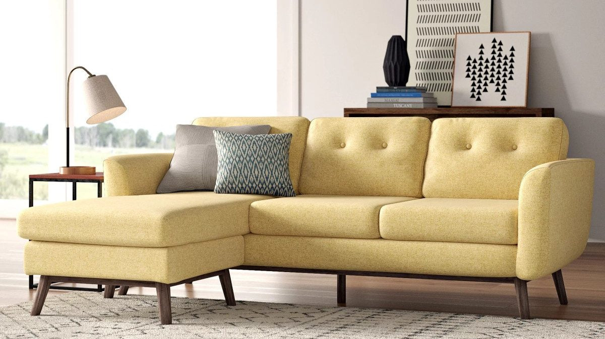 A modern living room with a yellow sectional sofa, featuring button-tufted back cushions and wooden legs. The sofa is adorned with two pillows, one gray and one patterned. A side table with a lamp is on the left, and a wooden console with decor is behind the sofa.