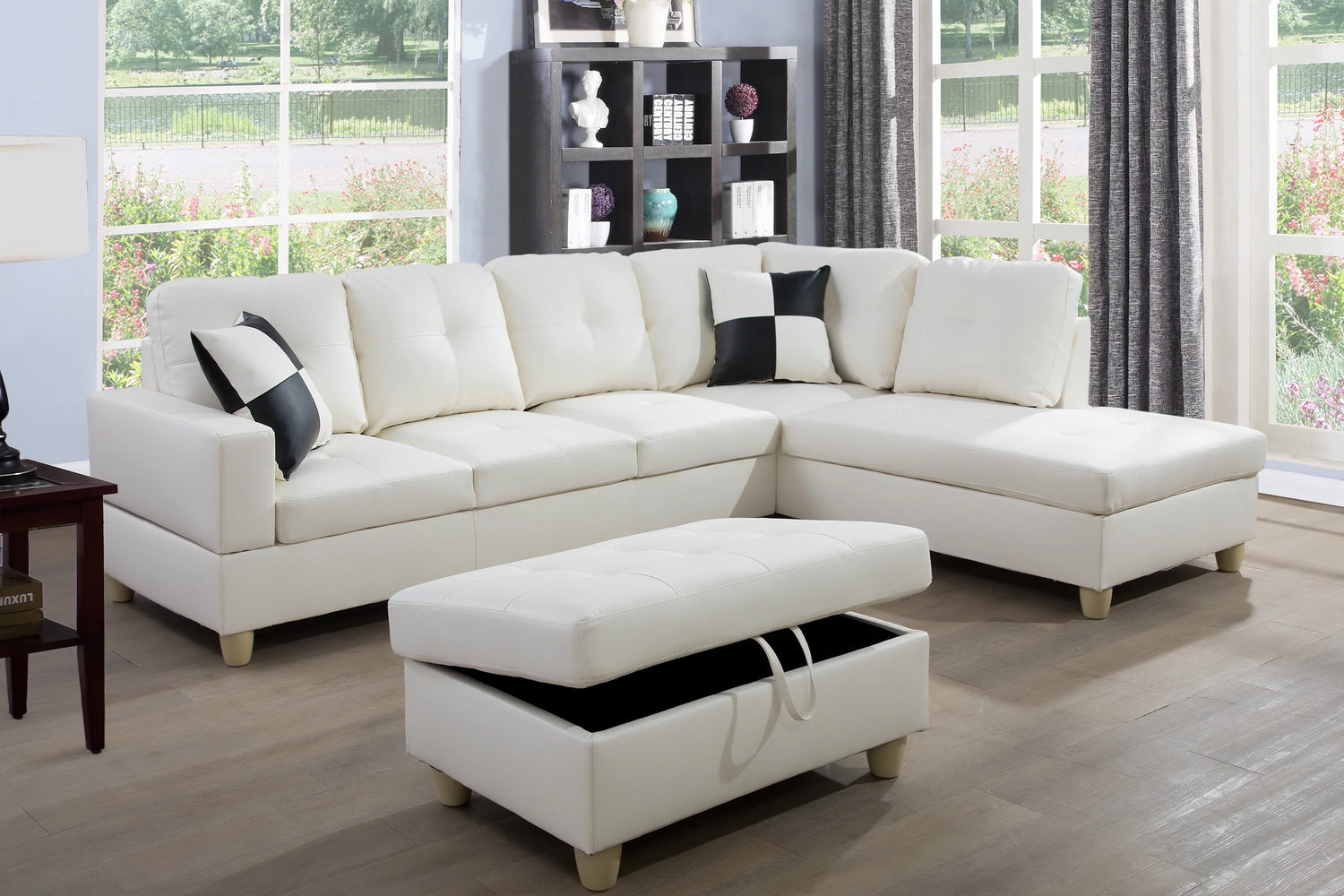 A spacious, bright living room features a white sectional sofa with black accent pillows. In the foreground, there is a matching white ottoman with storage and an open lid. Large windows with gray curtains allow natural light to flood the room, highlighting wooden floors.