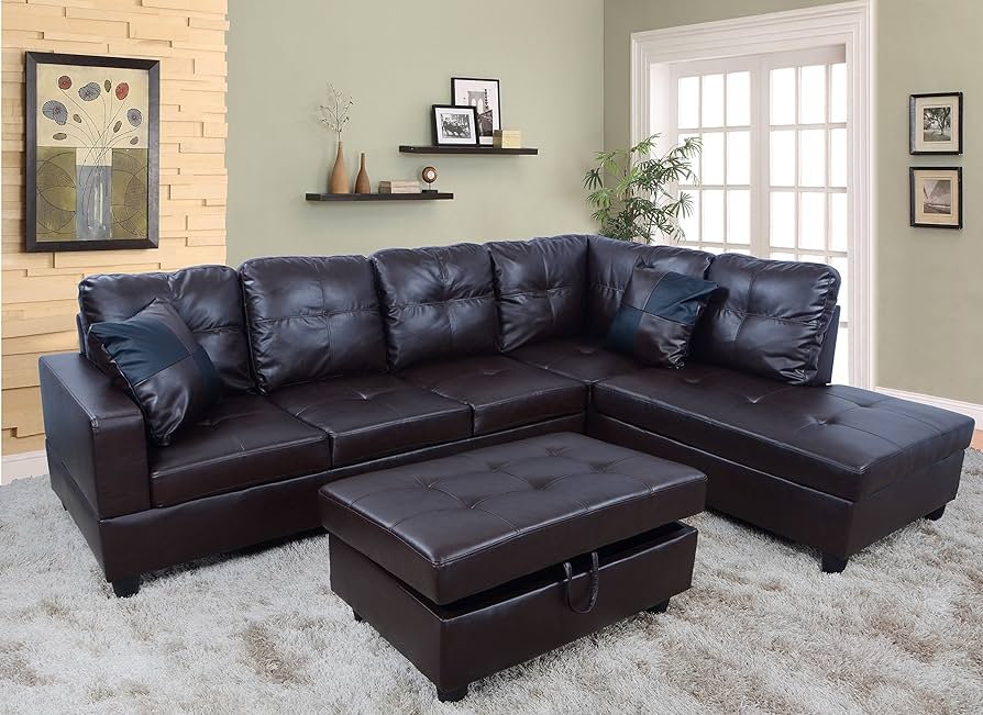 A cozy living room features a dark brown leather sectional couch with two blue accent pillows, accompanied by a matching tufted ottoman. The room has light green walls adorned with framed art and shelves, and is complemented by a plush light gray rug and a potted plant.