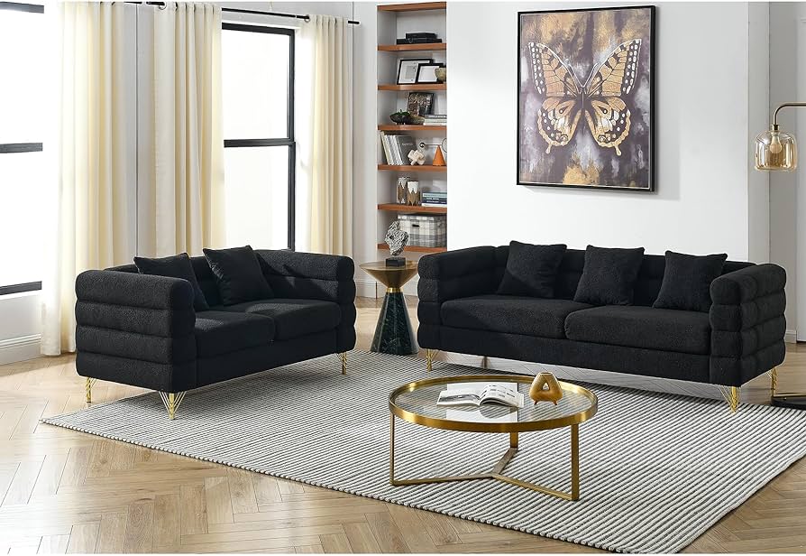 A modern living room with two black sofas, a round coffee table, and a large butterfly artwork on the wall. The room has beige curtains, a striped rug, and a bookshelf in the corner. The table has a small black tray, a glass with a candle, and a yellow ornament.
