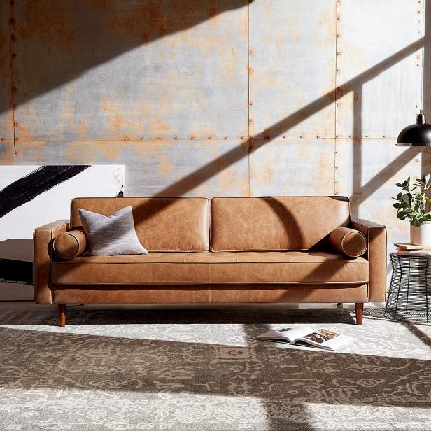 A sleek, mid-century modern tan leather sofa with square arms and a pair of round bolster pillows sits against a textured wall. A patterned area rug, a gray throw pillow, a potted plant, and a floor lamp decorate the space. Shadows from windows create geometric shapes on the wall.