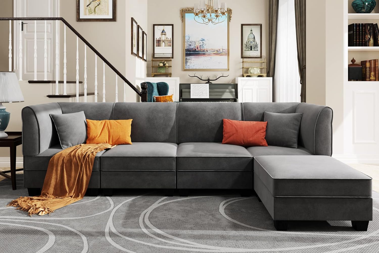 A modern living room features a gray sectional sofa adorned with orange and gray throw pillows. A mustard-yellow throw blanket is draped over one end. The background includes framed artwork, a staircase with a white railing, built-in shelves, and elegant decor.