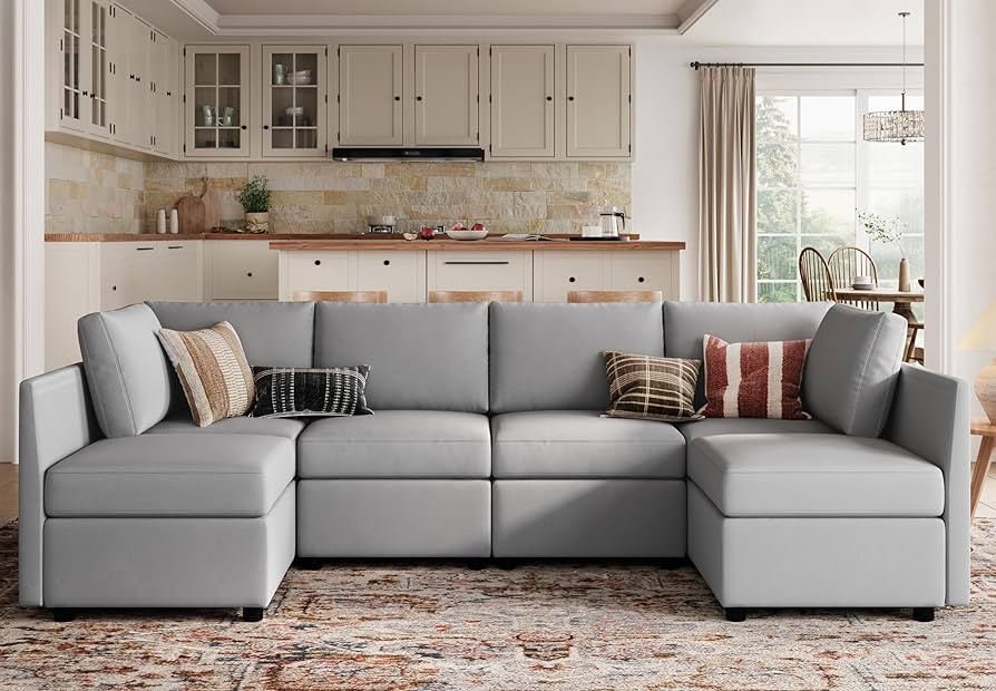 A modern living room with an L-shaped gray sectional sofa adorned with assorted throw pillows. Behind the sofa is a kitchen with cream-colored cabinets and a backsplash with beige tiles. Large windows allow light to flood in, highlighting the cozy space.