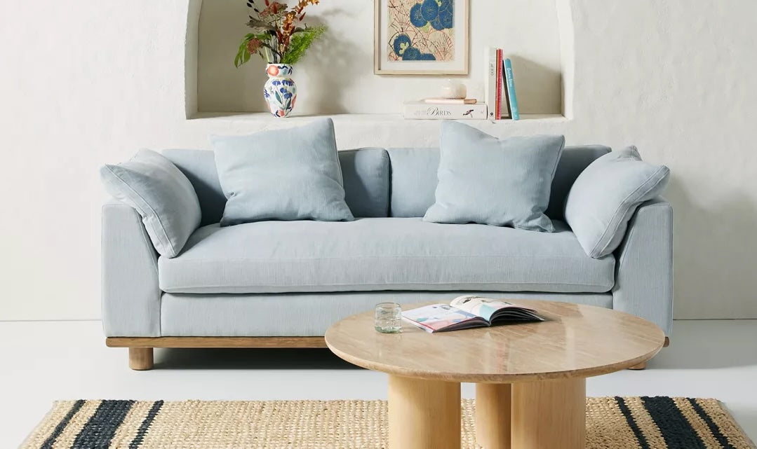 A light blue sofa with four matching cushions is set against a white wall with a decorative alcove. Above the sofa, there's a framed picture, a vase with flowers, and some books. In front of the sofa is a round wooden coffee table with an open magazine and a glass.