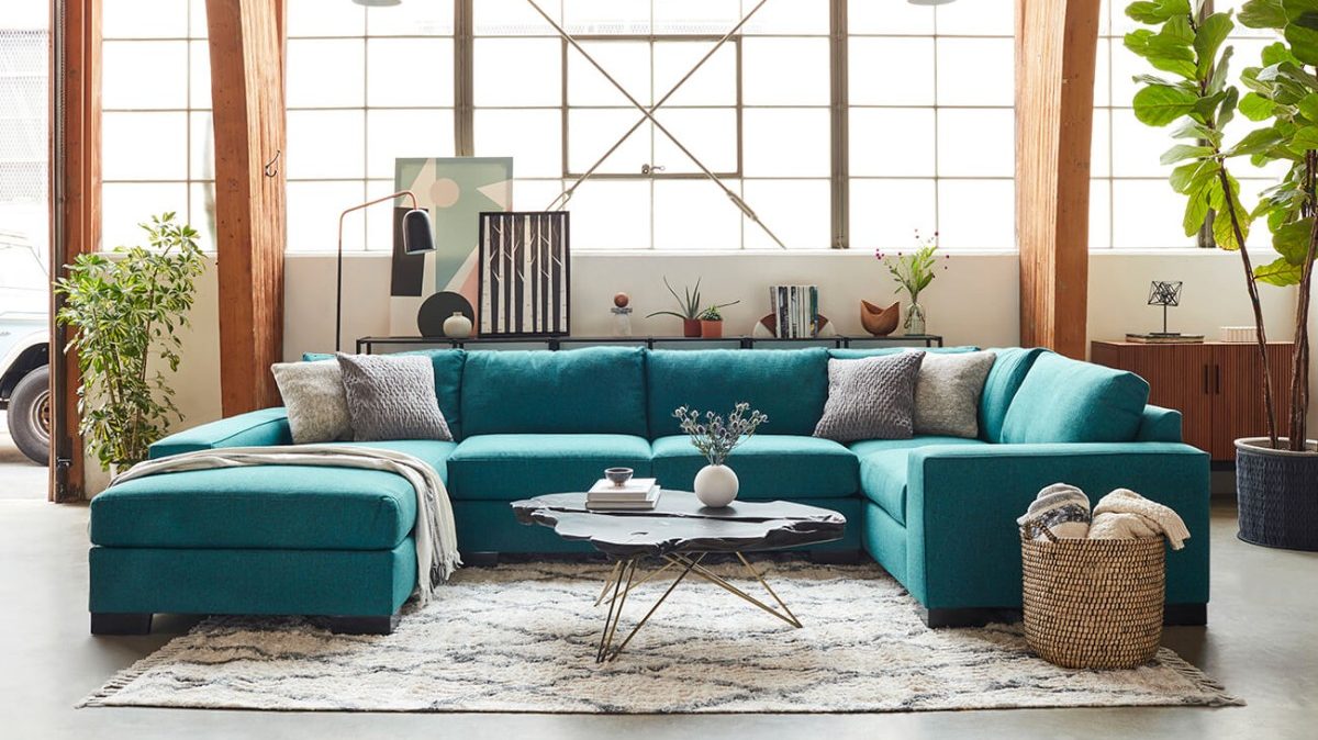 Bright and spacious living room with a large teal sectional sofa. The sofa has gray and white throw pillows, and a black modern coffee table is placed on a patterned rug. The room features large windows, plants, and various decorative items.