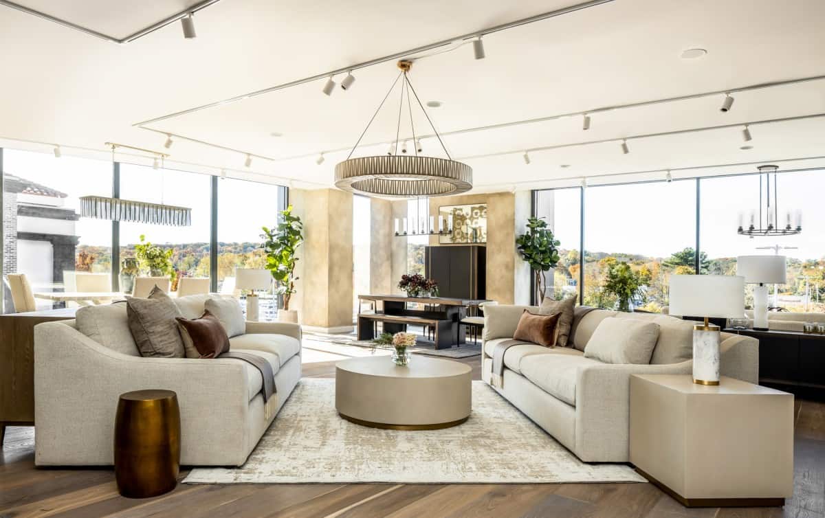 A spacious and modern living room features two beige sofas with brown and white cushions, a large circular coffee table, and a distinctive chandelier. Floor-to-ceiling windows offer a panoramic view of a landscape with autumn foliage. Decor includes plants and candles.