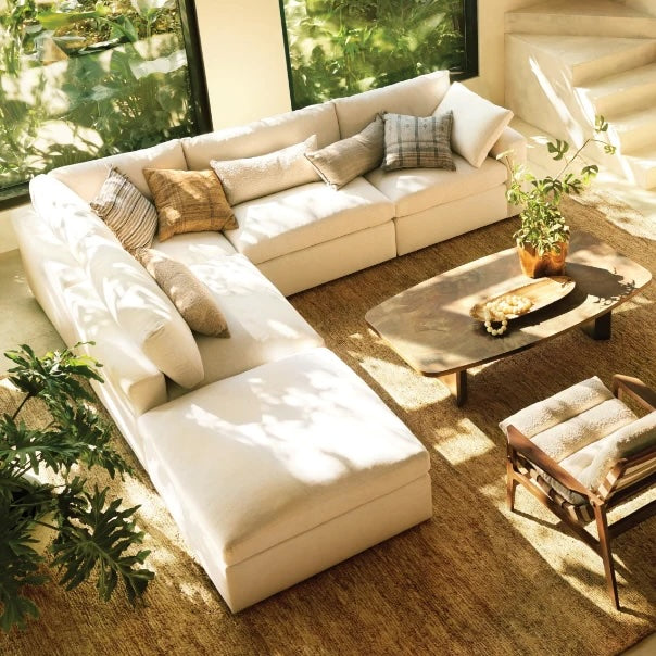 A bright living room features a white L-shaped sofa adorned with cushions, a wooden coffee table with decorative items, and a wooden armchair with a cushion. Large windows reveal lush greenery outside, and sunlight streams in, casting shadows on the beige carpet.