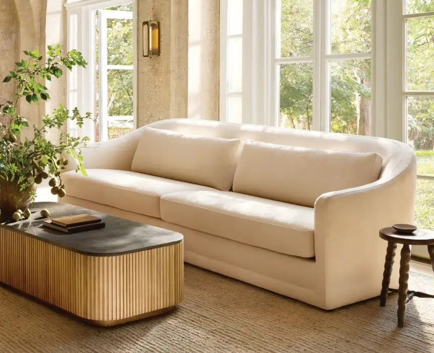 A modern living room with large windows bringing in natural light, featuring a cream-colored sofa with two cushions, a rectangular coffee table with a black top and ribbed sides, a small round side table, and a leafy green plant.