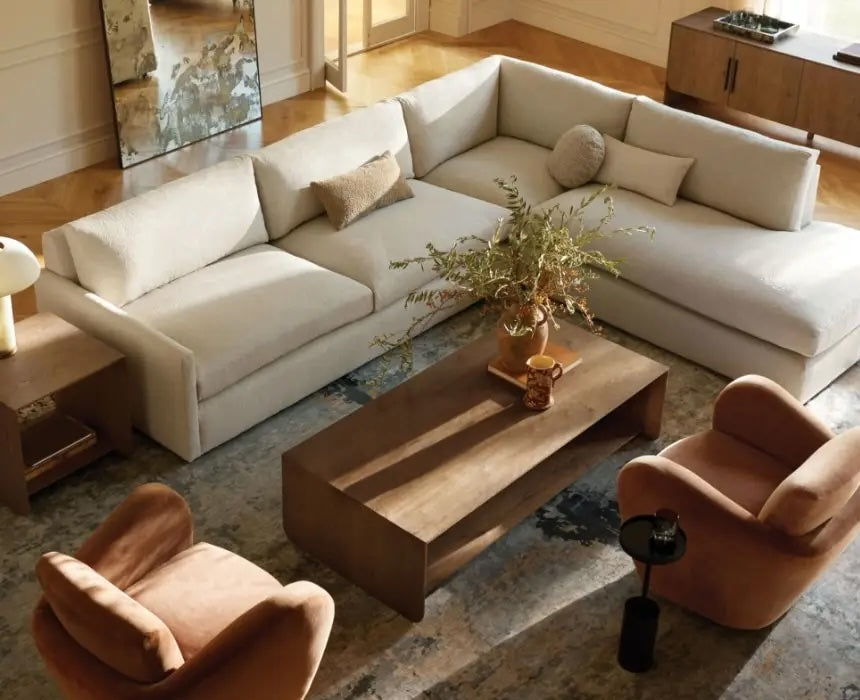 A modern living room features a large beige sectional sofa with several throw pillows. A wooden coffee table is centered on a patterned rug, flanked by two cushioned brown chairs. A side table with a lamp and a sideboard with decor complete the space.