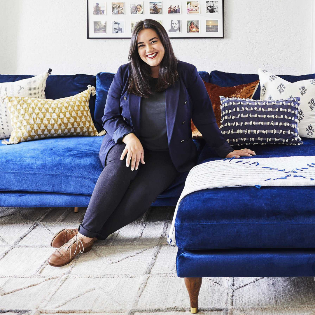 A person with long dark hair is sitting on a blue sectional sofa, smiling. They are wearing a dark blazer, dark pants, and brown shoes. The sofa is adorned with various patterned pillows. A photo collage is hung on the white wall behind the sofa.