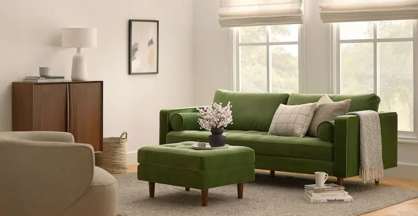 A cozy living room with a green couch, matching green ottoman, and a beige armchair. The couch is adorned with pillows, and a vase of pink flowers sits on the ottoman. Large windows with neutral curtains, a wooden cabinet, and a framed picture complete the room's decor.