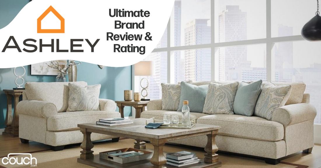A cozy living room featuring Ashley Furniture with a beige sofa and loveseat, both adorned with blue and beige pillows. A wooden coffee table with decorative items sits in front of the furniture. Large windows reveal a cityscape background. Text reads "Ashley: Ultimate Brand Review & Rating".