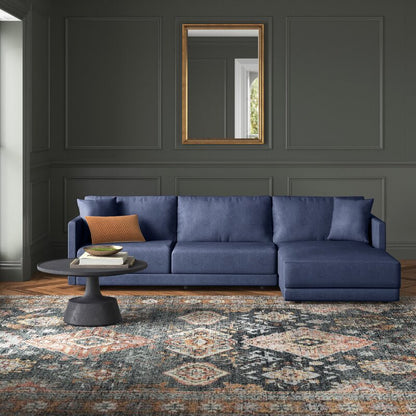 A spacious living room with dark green walls features the Astra 2-Piece Upholstered Sectional in a large blue design, complemented by a patterned area rug. A modern coffee table holds two decorative objects, while a gold-framed mirror hangs above the sectional, reflecting the window's light.