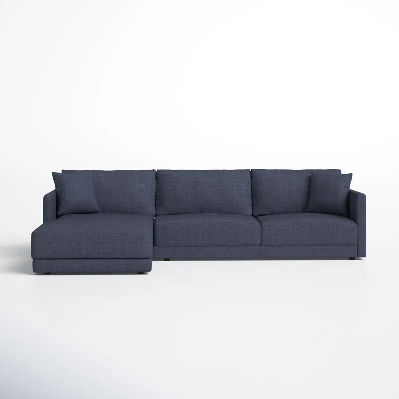 The Astra 2-Piece Upholstered Sectional is a contemporary dark gray L-shaped sofa featuring three seat cushions and a chaise lounge on the left side, set against a plain white wall and floor.