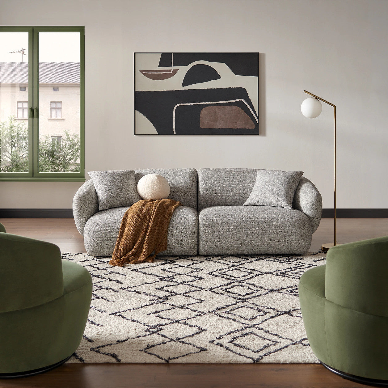 A modern living room featuring the Auburn Performance Fabric Sofa adorned with a brown throw, complemented by two green swivel chairs and a geometric black-and-white rug. Beside the sofa stands a floor lamp with a round shade, and an abstract painting is displayed on the wall.