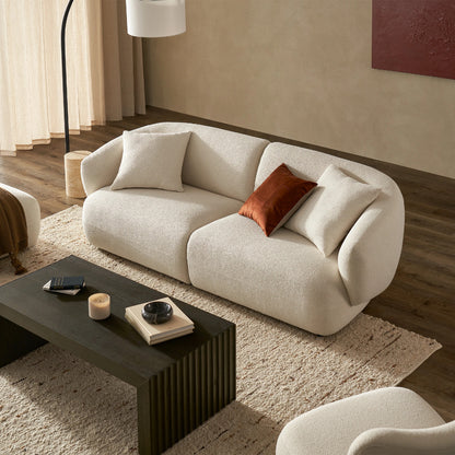 A modern living room showcases the Auburn Performance Fabric Sofa, elegantly curved in white and adorned with white and brown pillows. It is paired with a dark wooden coffee table that holds a candle, a book, and a decorative item. The room features a textured beige rug on the floor and neutral walls accented by abstract art.