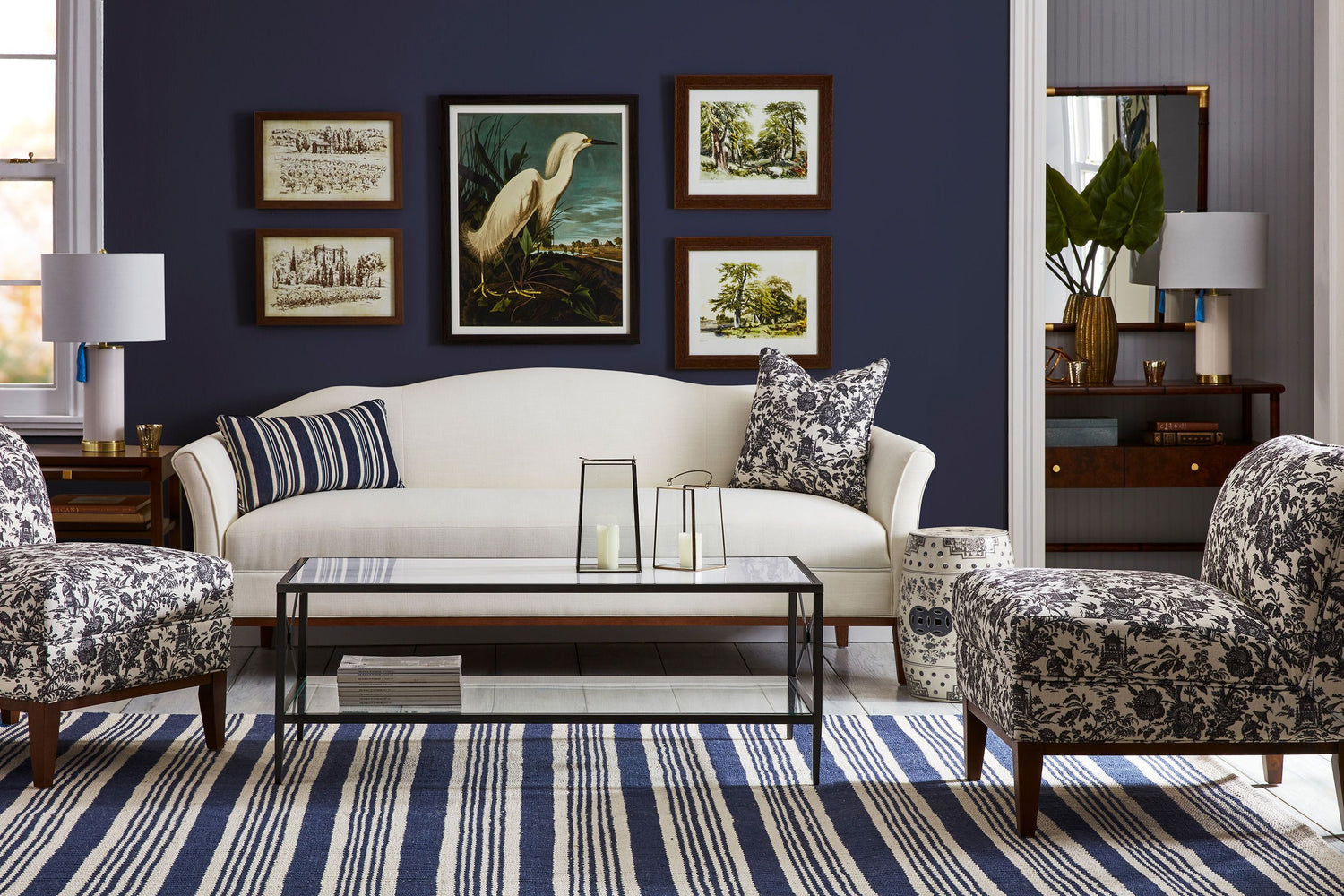 A stylish living room with a white sofa, flanked by two blue-white patterned chairs. A glass coffee table sits on a blue and white striped rug. Navy blue walls feature framed art, including a bird portrait. Lamps and decorative accents complete the look.