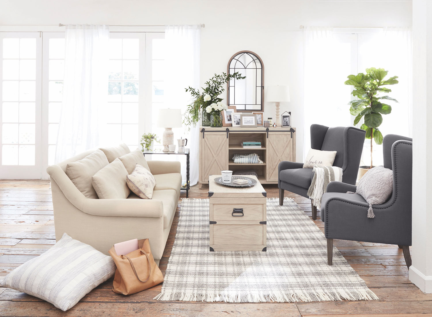 A well-lit, modern living room with a beige sofa, two gray armchairs, a wooden coffee table, and a patterned rug. Decor includes potted plants, a wall mirror, and various cushions. A leather bag and striped pillow are on the floor, and the room features large windows with white curtains.