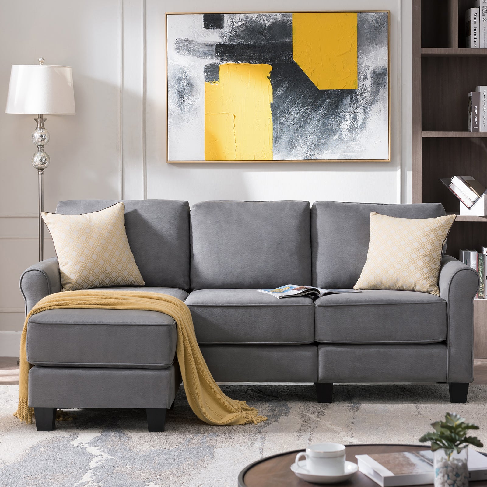 A cozy living room featuring a gray sofa with a chaise lounge, adorned with two yellow-patterned pillows and a yellow throw blanket. Behind the sofa is a modern abstract painting with yellow, black, and gray elements. A lamp and shelving unit flank the setup.