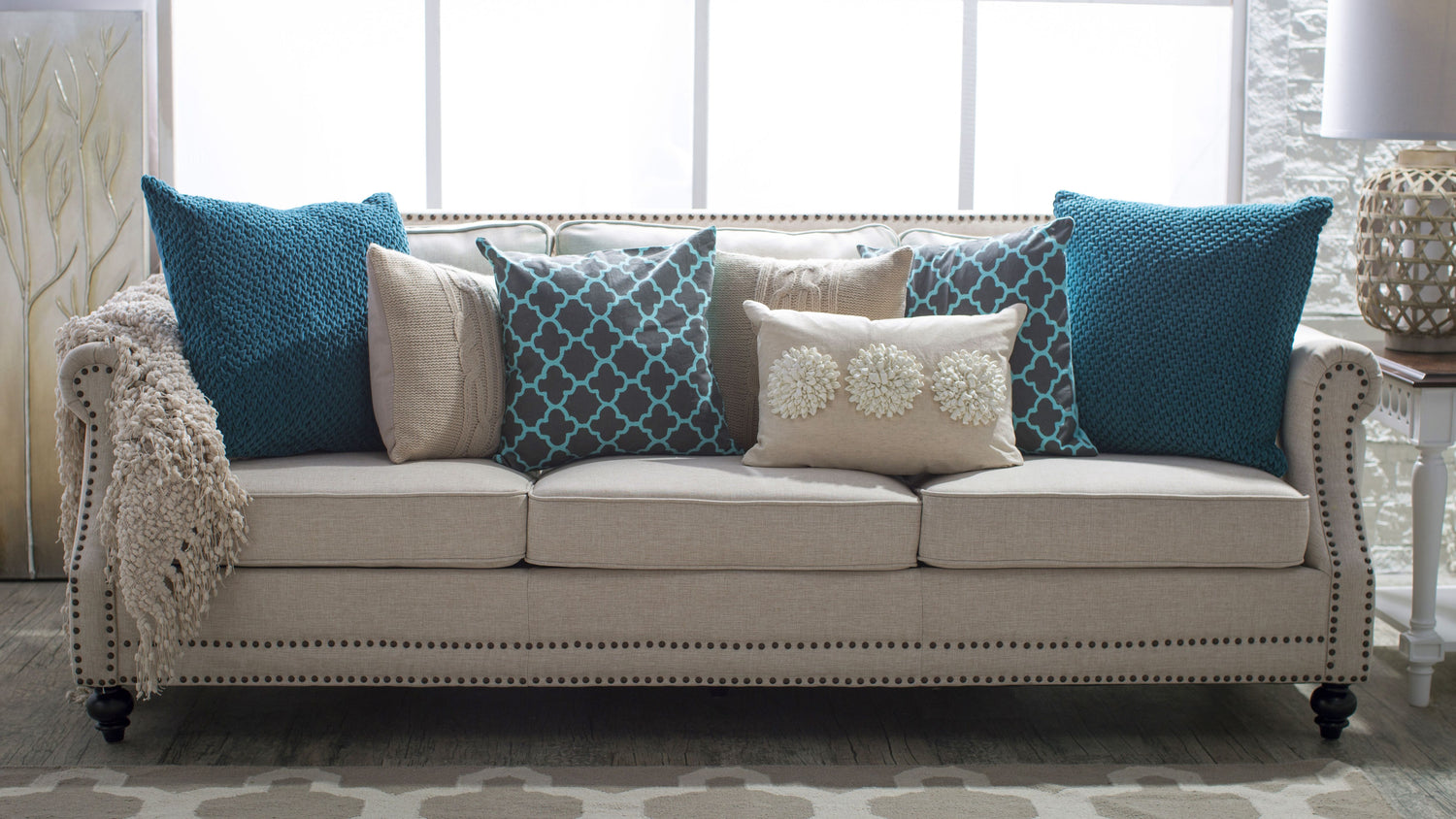 A beige sofa adorned with nailhead trim is decorated with an assortment of throw pillows in shades of teal and beige. The pillows feature various textures and patterns, including solid knits, geometric designs, and floral accents. A knit throw is draped on one armrest.
