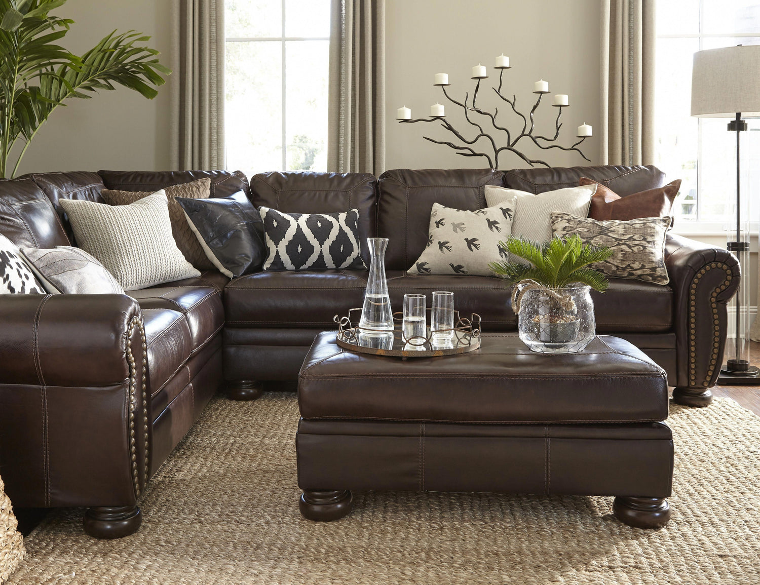 A cozy living room with a dark brown leather sectional sofa adorned with patterned throw pillows. A matching leather ottoman holds a decorative tray with glassware and a potted plant. A candelabra stands behind the sofa, with large windows and light curtains in the background.