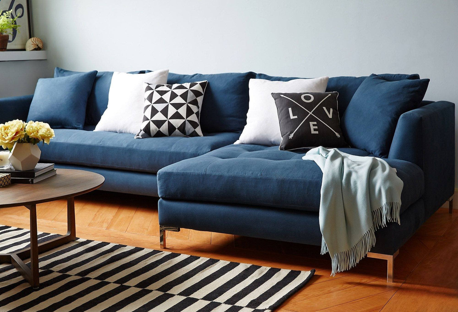 A modern living room with a blue sectional sofa adorned with decorative pillows, including a geometric design and one with the word "LOVE". A light blue throw blanket is draped over the sofa. The room features a wooden floor, round coffee table, and a black-and-white striped rug.