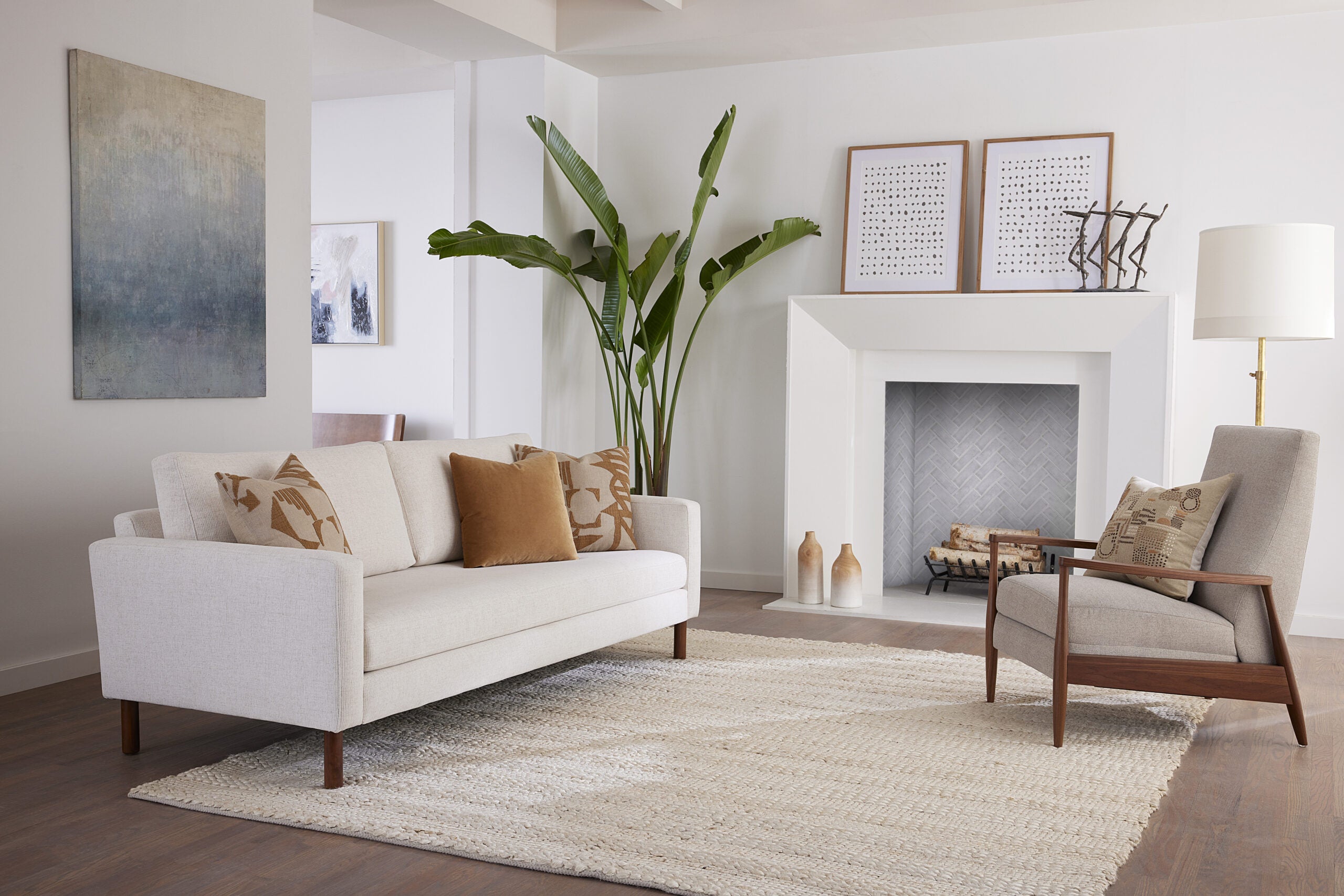A modern living room features a beige sofa with decorative cushions, an armchair, a large indoor plant, and minimalist art on the walls. The room has a light, neutral color palette with wooden accents and a textured area rug on the floor.