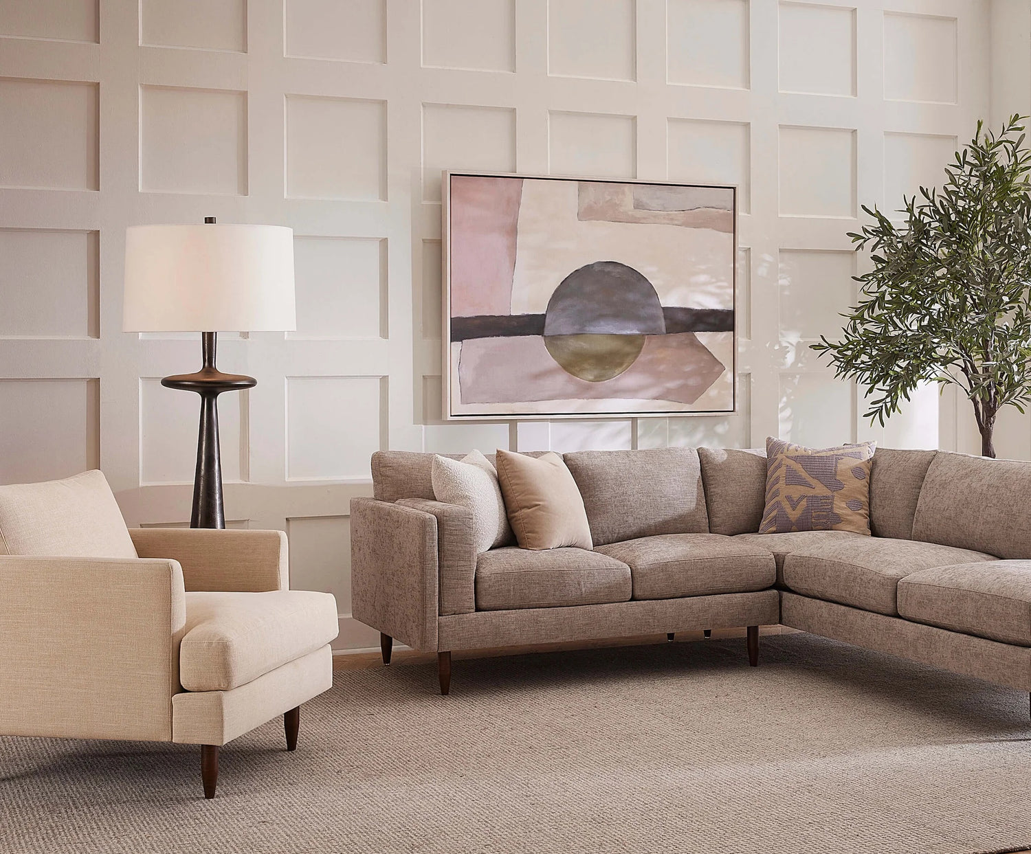 A modern living room with a light beige sectional sofa, a matching armchair, and a tall floor lamp. The walls feature a grid-like panel design and a large abstract painting. An artificial tree adds a touch of greenery. The space is carpeted in a neutral tone.