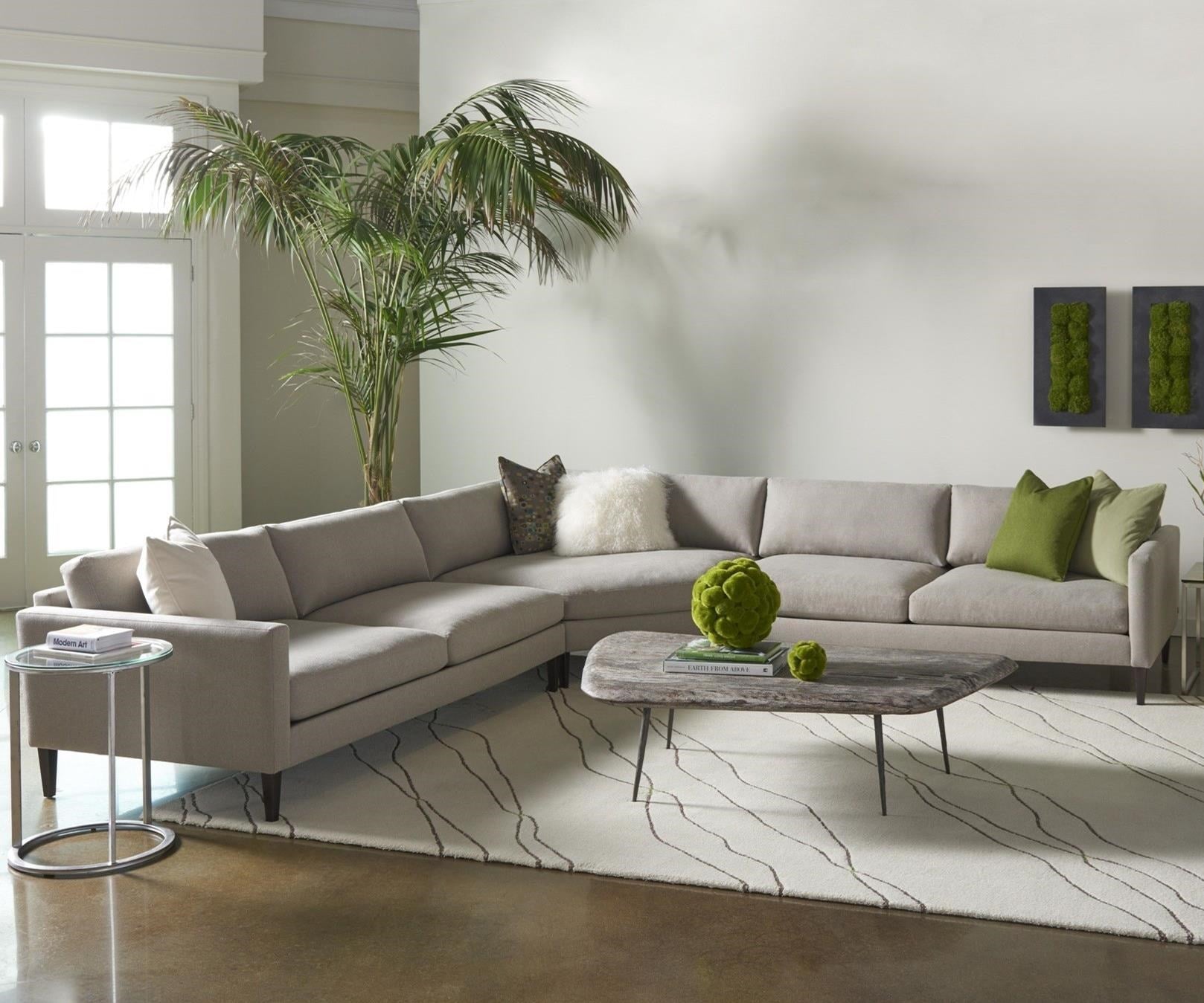 A modern living room features a light-gray sectional sofa adorned with green and white throw pillows. A wooden coffee table holds a decorative green sphere. Potted palm trees and framed green art add a touch of nature. The room has a rug with wavy lines and a side table.