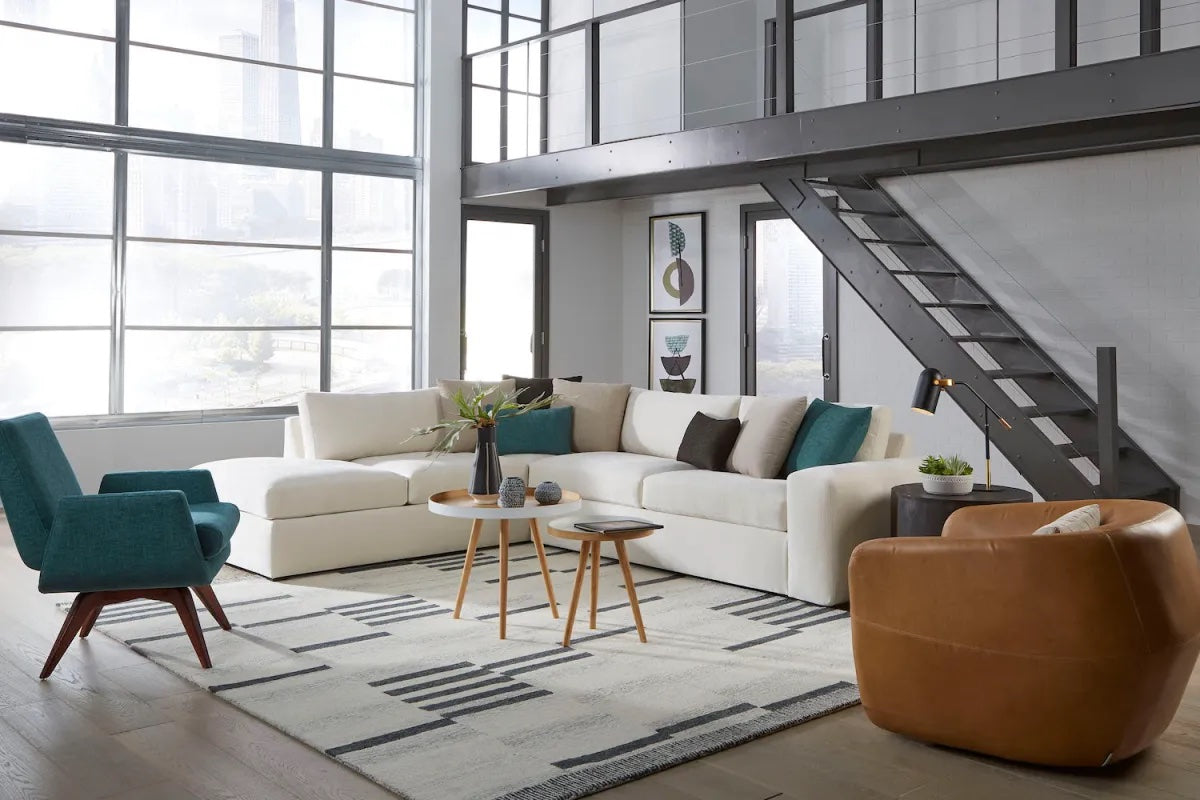 A modern loft living room featuring a large L-shaped white sofa adorned with throw pillows. There are teal and brown accent chairs, a pair of round wooden coffee tables with vases, a patterned rug, industrial staircase, and large windows with ample natural light.