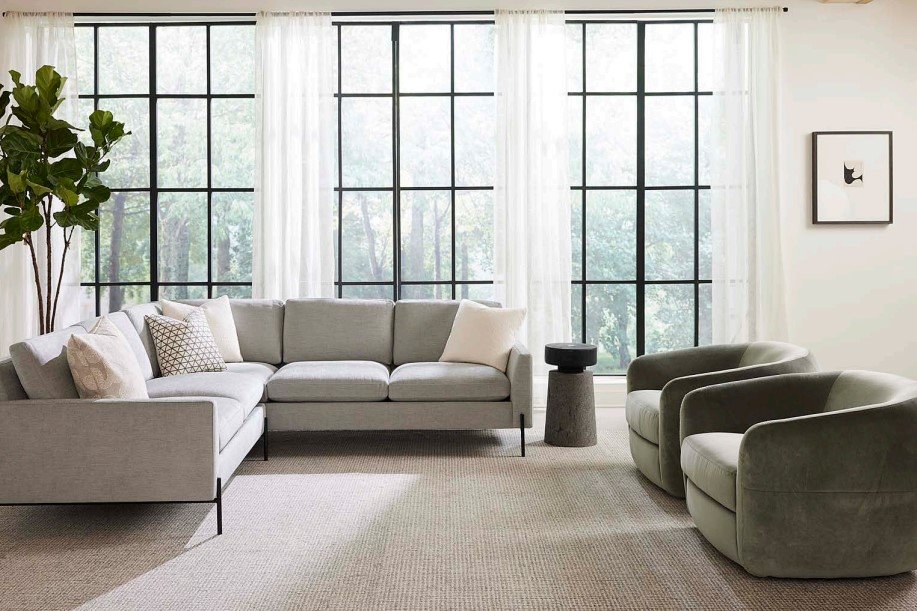 A modern living room with large windows covered by sheer white curtains. A light gray sectional sofa with decorative pillows is arranged beside a small round side table. Two gray armchairs face the sofa, and a tall potted plant and framed artwork complete the scene.