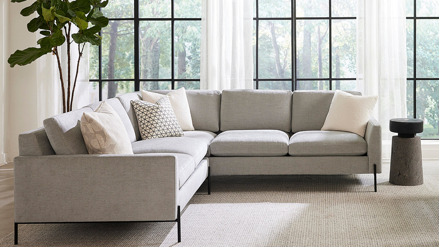 A modern living room with a large gray sectional sofa adorned with white and patterned throw pillows. The room features large windows with sheer white curtains, revealing a green outdoor scene. A tall potted plant stands in the corner next to the sofa.